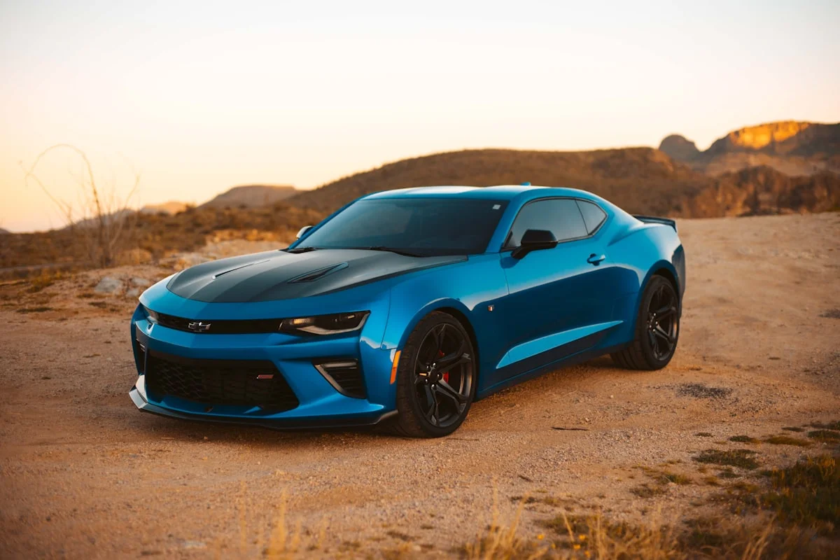 Blue Chevrolet Camaro coupé parked on a desert track at sunset, with red rock hills behind.