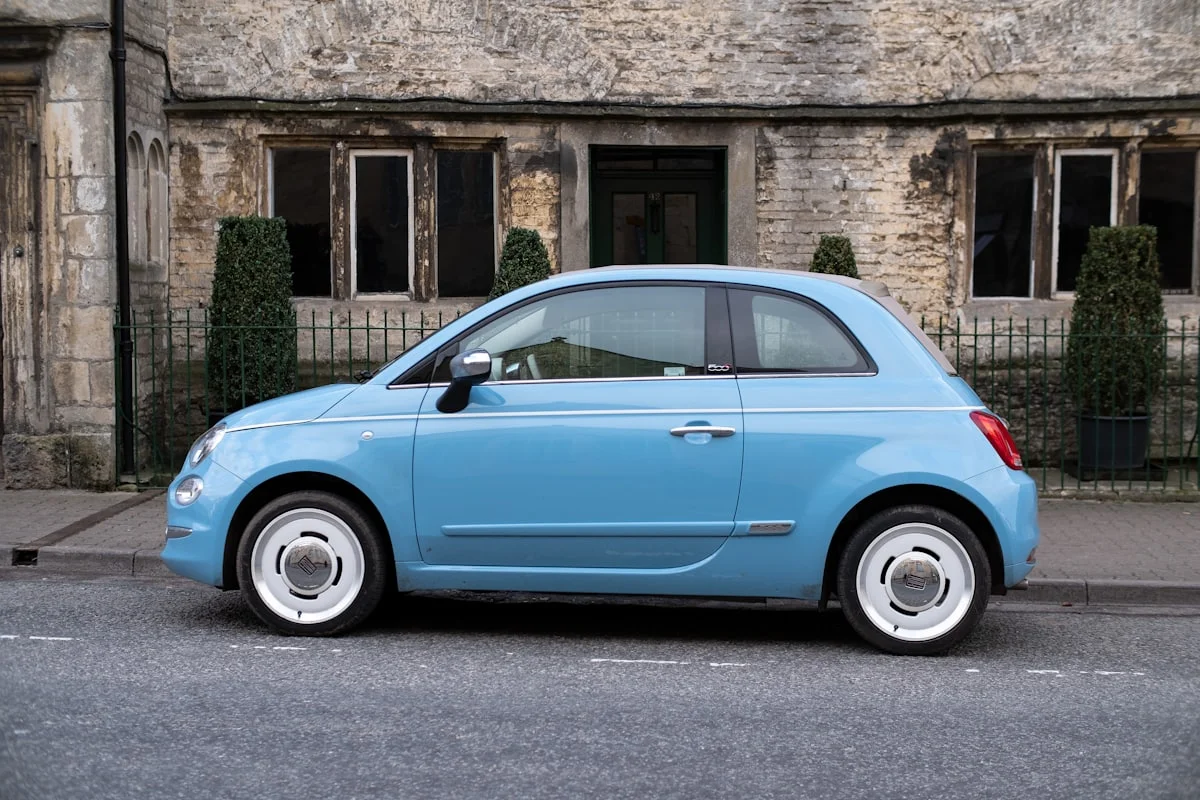 Light blue Fiat 500 city car parked in profile against a honey-coloured stone building.
