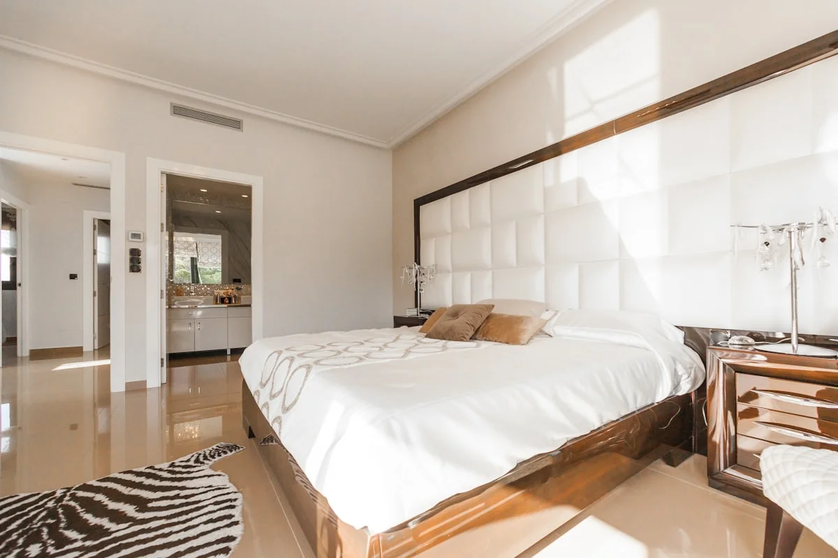 Bright bedroom with a tall padded white headboard, a polished timber bedframe and an open door to a marble bathroom.