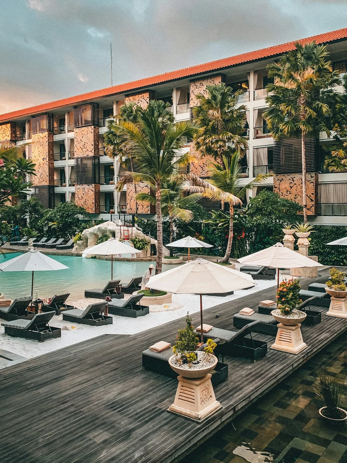 Resort wing with a terracotta-tiled roof and carved wall panels above a sand-edged pool, palms and a timber sun deck.