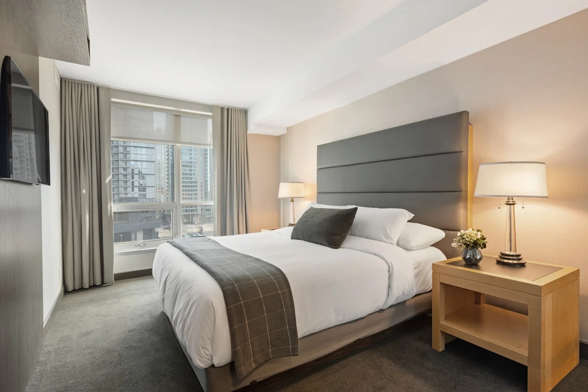 Bedroom with a grey padded headboard, twin bedside lamps and a high-rise city view through the window.