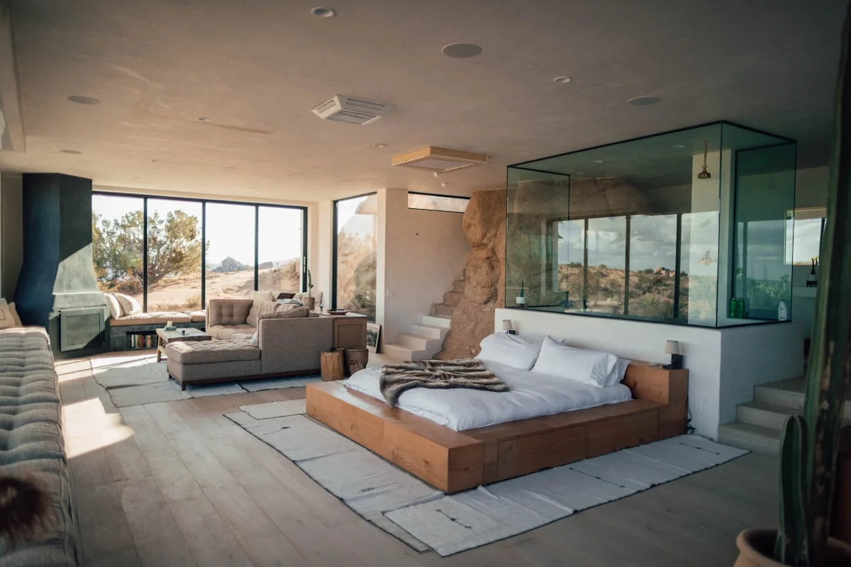 Open-plan room with a low timber platform bed, a glass-walled bathroom and windows onto boulders outside.